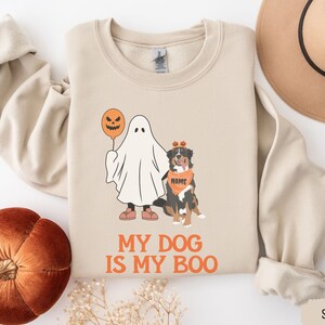 May include: A beige sweatshirt with a graphic of a dog dressed as a ghost holding a jack-o-lantern. The text "My Dog Is My Boo" is printed below the graphic. The sweatshirt is available in the color "Sand".