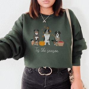 May include: A forest green sweatshirt with a graphic of three Bernese Mountain Dogs wearing fall-themed accessories. The text "'Tis the season" is printed below the dogs.