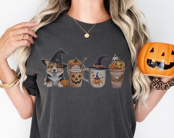 May include: A dark gray t-shirt with a graphic of a corgi wearing a witch hat and holding a pumpkin. The graphic also includes four mugs with Halloween-themed designs. The t-shirt is being held by a person who is also holding a pumpkin-shaped mug.