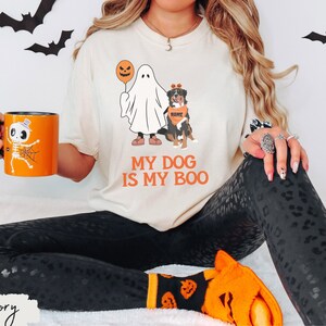 May include: Ivory-colored t-shirt with the text "MY DOG IS MY BOO" in orange. The shirt features a ghost holding a balloon and a dog wearing a bandana. An orange mug with a skeleton design is also visible.