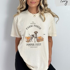 May include: A cream colored t-shirt with a graphic of a dog standing in front of pumpkins. The graphic says "The Original Farm Fresh Est'd 1990 Pumpkin Patch Open Daily".