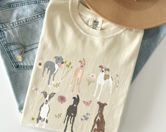 May include: Ivory-colored t-shirt with a crew neck featuring a graphic of various dog breeds with floral accents. The shirt is paired with light-wash denim jeans and a beige hat. A delicate necklace is also visible.