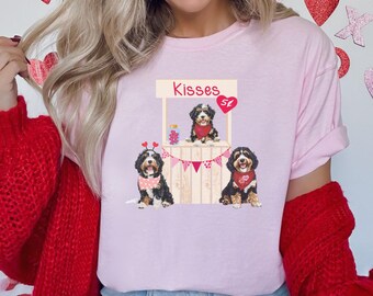 May include: Pink t-shirt with a graphic of three dogs standing in front of a Valentine's Day themed stand that says "Kisses St."