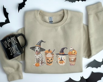 May include: Sand-colored sweatshirt featuring a Dalmatian dog wearing a witch hat, alongside pumpkin spice latte-style mugs with Halloween designs. A black mug with "VAMPIRE JUICE" text and paper bats complete the festive theme.