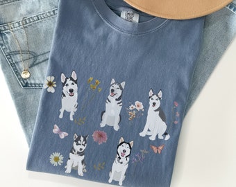 Può includere: Una maglietta blu con una grafica di cinque Husky siberiani con accenti floreali. La maglietta ha il testo "COMFY COLORS" sull'etichetta.