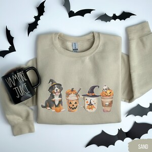 May include: Sand-colored sweatshirt with a Halloween-themed graphic featuring a dog in a witch hat, pumpkin spice latte cups, and a black mug that says "VAMPIRE JUICE". Black bat cutouts are scattered around the sweatshirt.