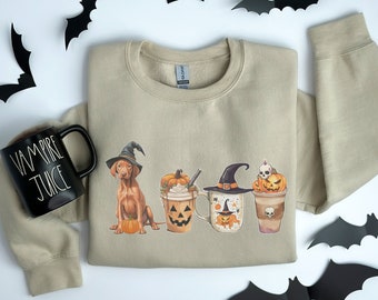 Può includere: Una felpa color beige con una grafica di un cane che indossa un cappello da strega, zucche e tazze da caffè a tema Halloween. Il testo "Vampire Juice" è su una tazza nera nell'immagine.