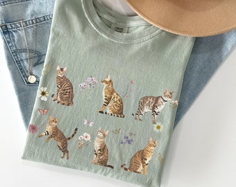 以下が含まれることがあります： 薄い緑色のTシャツに、茶色と黒のベンガル猫が花と蝶と共に見られるプリントが施されています。シャツには「COMFORT」の文字が見えます。
