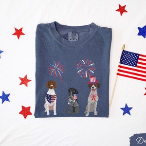 May include: A navy blue t-shirt with three dogs wearing red, white, and blue patriotic accessories. The dogs are surrounded by fireworks. The shirt has a tag that says "Comfort Colors" and "M/M/M".