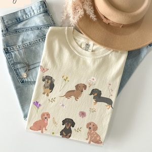 以下が含まれることがあります： アイボリーカラーのTシャツに、複数のダックスフントの犬と花のアクセントが描かれたグラフィックがプリントされています。Tシャツのタグには「COMFORT COLORS」の文字が見えます。
