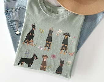 May include: A light green t-shirt with a graphic print of five Doberman Pinschers with flowers and butterflies. The shirt is laid out on a light blue denim background.