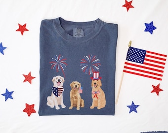 Puede incluir: Una camiseta azul marino con tres perros golden retriever que llevan accesorios patrióticos. Los perros están de pie frente a un espectáculo de fuegos artificiales. La camiseta tiene el texto "Live Free" en la etiqueta.