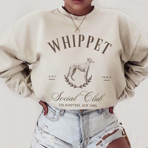Puede incluir: Una sudadera beige con el texto "Whippet Social Club" y un gr&aacute;fico de un perro whippet. El texto "Est. 1983" est&aacute; debajo del gr&aacute;fico. La sudadera la lleva puesta una persona.