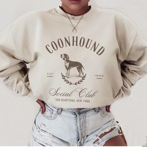 May include: A beige sweatshirt with a brown graphic print of a dog and the text "COONHOUND Social Club THE HAMPTONS, NEW YORK"