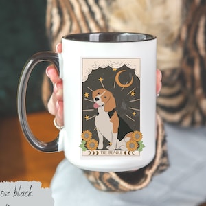 May include: A white ceramic mug with a black handle and a black rim. The mug features a graphic of a beagle dog sitting in front of a crescent moon and stars. The text "THE BEAGLE" is printed below the dog. The mug is 15 ounces.