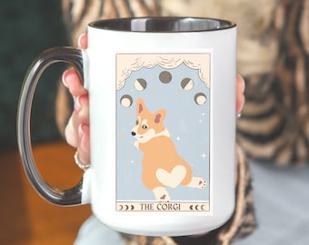 May include: A white 15 oz mug with a black handle and interior. The mug features a whimsical illustration of a corgi dog with a heart-shaped marking, set against a light blue background with moon phases. Text reads "THE CORGI".