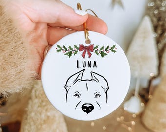 May include: White ceramic Christmas ornament featuring a black line drawing of a dog's face. The word "LUNA" is above the dog's face. The ornament is decorated with a red bow and green holly.