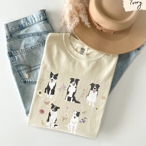 May include: A cream colored t-shirt with a graphic of five black and white border collies with floral accents. The shirt is laid out on a light blue denim background with a brown hat and a dried flower arrangement.