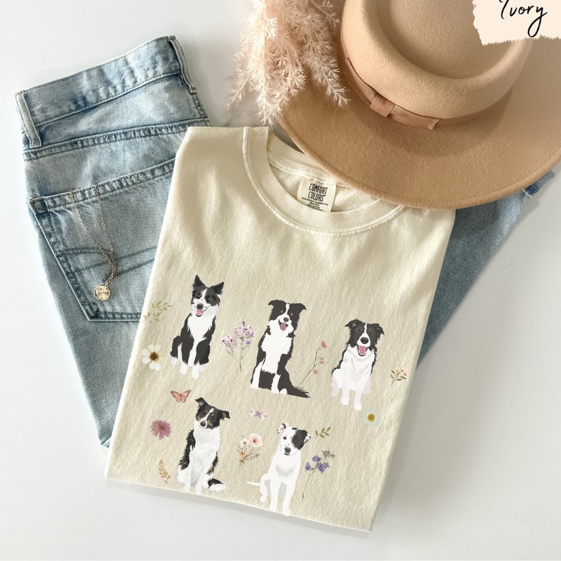 May include: A cream colored t-shirt with a graphic of five black and white border collies with floral accents. The shirt is laid out on a light blue denim background with a brown hat and a dried flower arrangement.