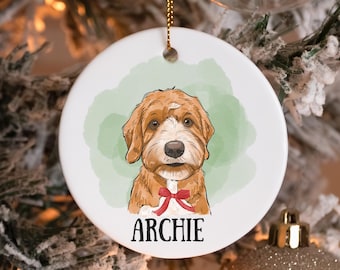 May include: A white, round Christmas ornament featuring a cartoon illustration of a brown and white dog with a red bow tie. The dog's name, "ARCHIE," is printed in black below the image. The ornament hangs from a gold string.