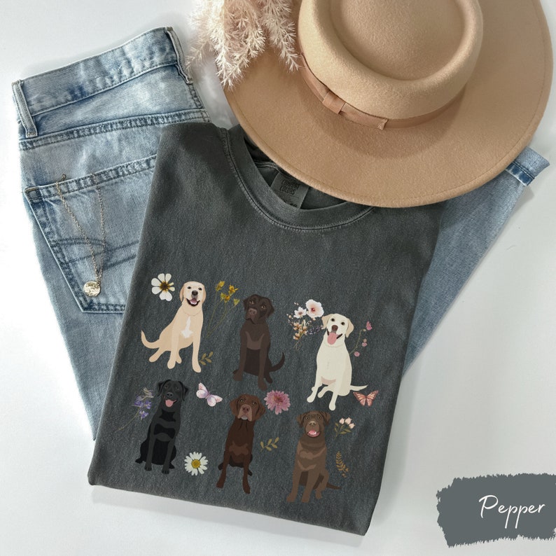 May include: A dark gray t-shirt with a graphic of six labrador retrievers in different colors, surrounded by flowers and butterflies. The text "Pepper" is visible in the bottom right corner.