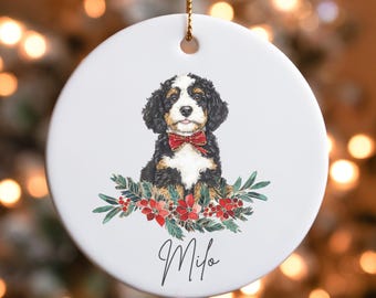 Puede incluir: Un adorno redondo de cerámica blanca con un perro negro, marrón y blanco con una pajarita roja. El perro está rodeado de hojas verdes y flores rojas. El adorno tiene el nombre "Milo" escrito en cursiva en la parte inferior.