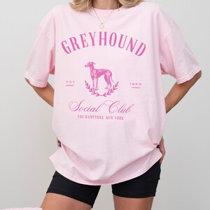 以下が含まれることがあります： ピンクのオーバーサイズTシャツに、グレーハウンドの犬のグラフィックプリントと「Greyhound Social Club The Hamptons, New York」のテキストがピンクでプリントされています。また、「Est 1983」のテキストもプリントされています。