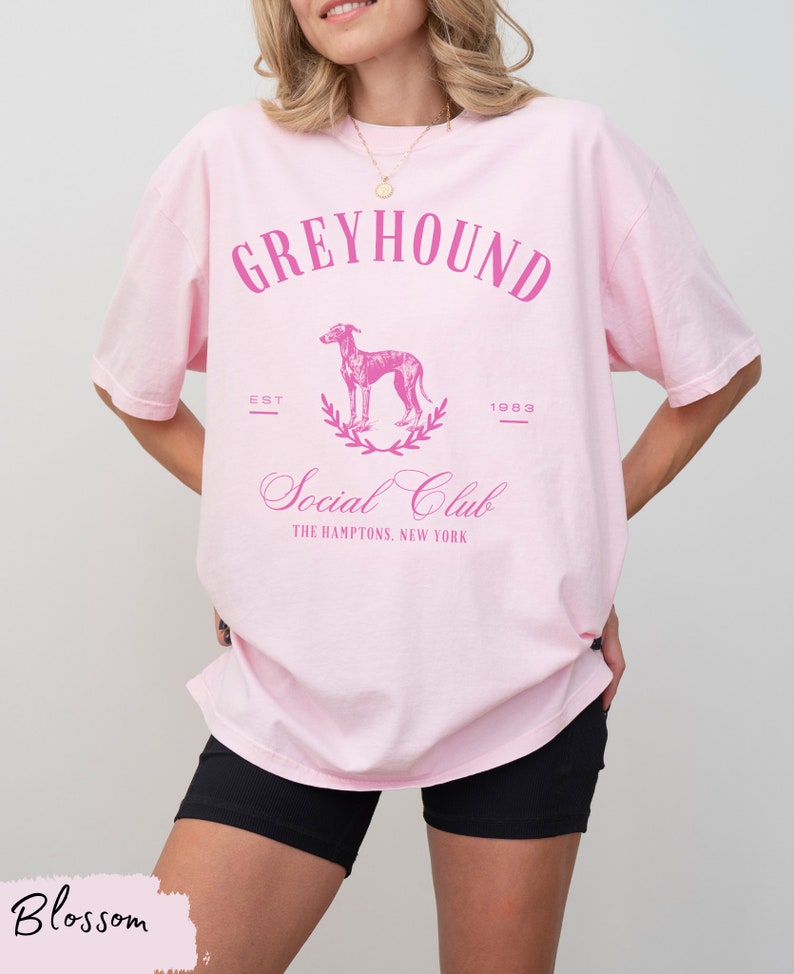 以下が含まれることがあります： ピンクのオーバーサイズTシャツに、グレーハウンドの犬のグラフィックプリントと「Greyhound Social Club The Hamptons, New York」のテキストがピンクでプリントされています。また、「Est 1983」のテキストもプリントされています。