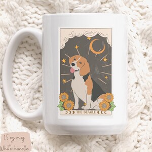 May include: White ceramic mug with a graphic of a beagle dog in a tarot card style illustration. The card has a dark brown background with stars, a crescent moon, and sunflowers. The text "THE BEAGLE" is printed below the dog.