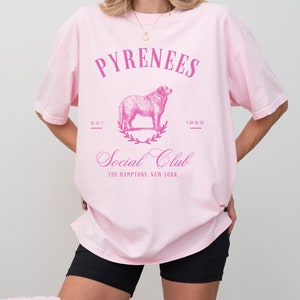 May include: Pink oversized t-shirt with a pink graphic of a dog, a wreath, and the text "Pyrenees Est 1989 Social Club The Hamptons, New York"