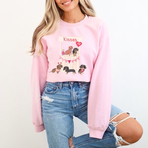 Puede incluir: Sudadera rosa con un gr&aacute;fico de una tienda de regalos de San Valent&iacute;n con tem&aacute;tica de dachshund con el texto "Kisses" sobre la tienda. La tienda tiene un toldo rosa y un letrero en forma de coraz&oacute;n.