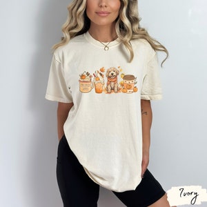 Op de afbeelding: Een wit T-shirt met een afbeelding van een hond die voor drie herfst-ge&iuml;nspireerde koffie-drankjes zit. De hond draagt een sjaal en er zijn pompoenen en bladeren rond de drankjes. De tekst op de mok is "Pumpkin Spice Latte".