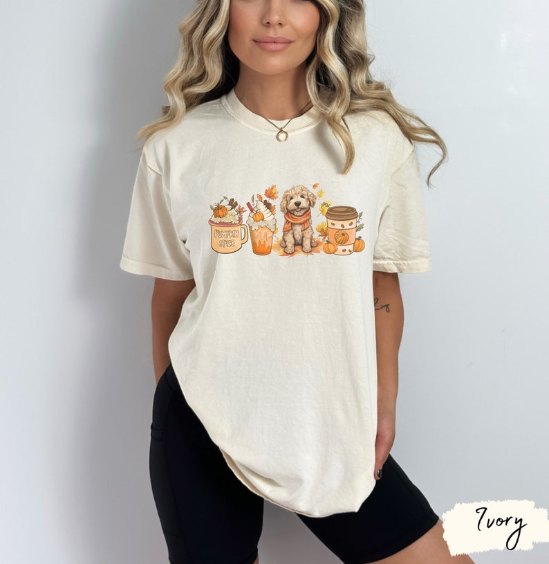 Op de afbeelding: Een wit T-shirt met een afbeelding van een hond die voor drie herfst-ge&iuml;nspireerde koffie-drankjes zit. De hond draagt een sjaal en er zijn pompoenen en bladeren rond de drankjes. De tekst op de mok is "Pumpkin Spice Latte".