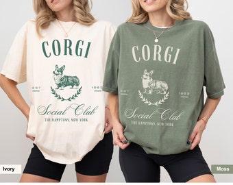 Può includere: Due donne indossano magliette oversize con una grafica vintage di un corgi. Il testo sulle magliette recita "CORGI Social Club The Hamptons, New York" con la data di fondazione "EST 1983". Le magliette sono disponibili in avorio e muschio.