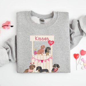 Puede incluir: Sudadera gris con un gr&aacute;fico de un puesto de besos de dachshund con el texto "Kisses 5&cent;". El gr&aacute;fico presenta cuatro dachshunds con corazones y una pancarta.