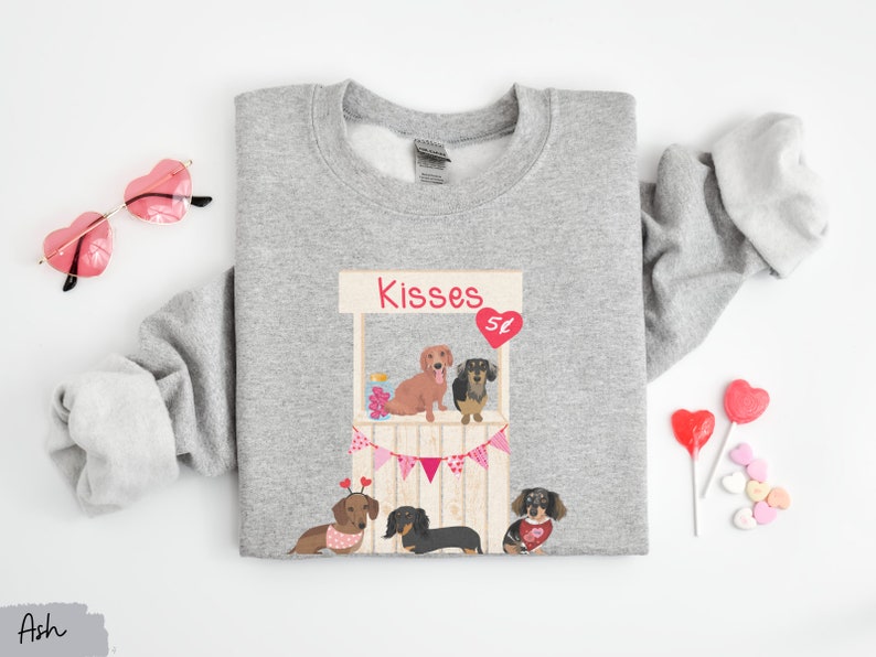 Puede incluir: Sudadera gris con un gr&aacute;fico de un puesto de besos de dachshund con el texto "Kisses 5&cent;". El gr&aacute;fico presenta cuatro dachshunds con corazones y una pancarta.