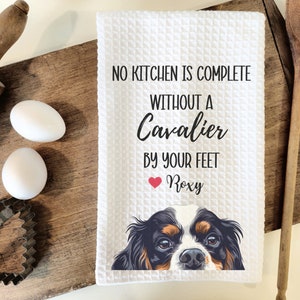 May include: White kitchen towel with the text "No kitchen is complete without a Cavalier by your feet" and a heart with the name "Roxy" below. A Cavalier King Charles Spaniel dog's face peeks out from the bottom of the towel.