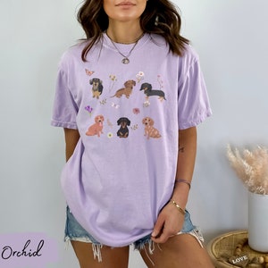 以下が含まれることがあります： ラベンダー色のTシャツに、花と蝶に囲まれた6匹のダックスフントのグラフィックがプリントされています。Tシャツはオーバーサイズでゆったりとしたフィット感です。