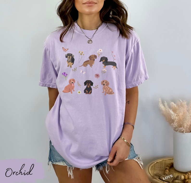 以下が含まれることがあります： ラベンダー色のTシャツに、花と蝶に囲まれた6匹のダックスフントのグラフィックがプリントされています。Tシャツはオーバーサイズでゆったりとしたフィット感です。