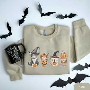 May include: A tan sweatshirt with a black cat wearing a witch's hat and holding a pumpkin. The cat is next to three mugs with pumpkin faces and Halloween decorations. The text "Vampire Juice" is on a black mug.