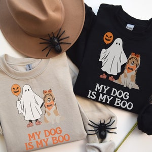 May include: Two sweatshirts, one beige and one black, feature a Halloween-themed graphic. The design includes a ghost holding a jack-o'-lantern balloon and a dog wearing a bandana. The text "MY DOG IS MY BOO" is printed below the graphic.