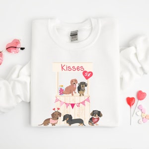 Puede incluir: Sudadera blanca con un gr&aacute;fico de tres perros salchicha en un puesto de besos tem&aacute;tico de San Valent&iacute;n. El texto "Kisses" est&aacute; sobre el puesto.