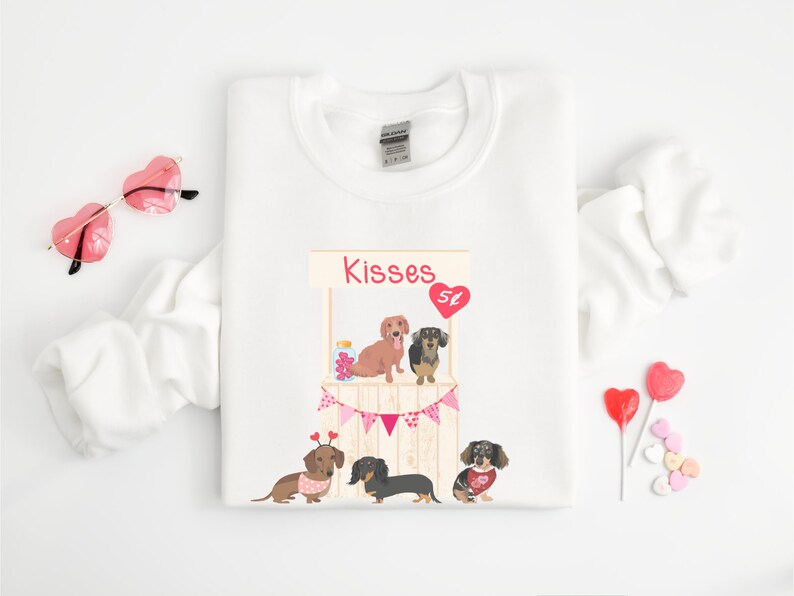 Puede incluir: Sudadera blanca con un gr&aacute;fico de tres perros salchicha en un puesto de besos tem&aacute;tico de San Valent&iacute;n. El texto "Kisses" est&aacute; sobre el puesto.