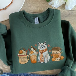 May include: A forest green sweatshirt with a cute cat wearing a scarf and surrounded by fall-themed drinks. The drinks include a pumpkin spice latte, a caramel latte, and a coffee cup with a pumpkin on it. The text on the pumpkin spice latte says "Pumpkin Spice".