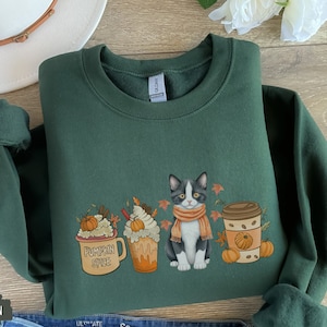 May include: Forest green sweatshirt featuring a fall-themed graphic. The design includes a cat wearing a scarf, pumpkin spice latte, and pumpkins. The sweatshirt is made of a soft material.