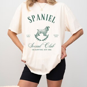 May include: A white t-shirt with a green graphic of a spaniel dog and the text "Spaniel Social Club, Est. 1983, The Hamptons, New York".