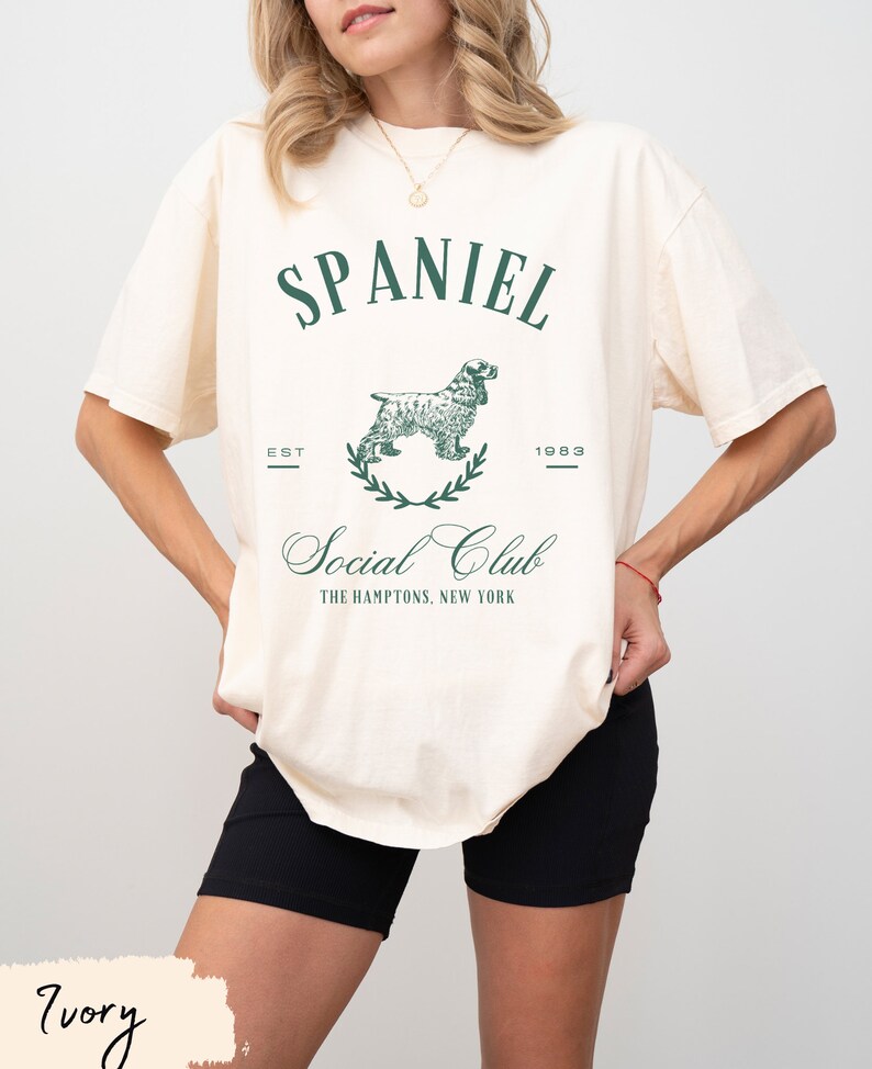 May include: A white t-shirt with a green graphic of a spaniel dog and the text "Spaniel Social Club, Est. 1983, The Hamptons, New York".