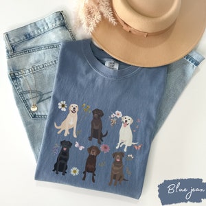 May include: A blue jean t-shirt with a graphic of five Labrador Retrievers in different colors, surrounded by flowers and butterflies. The text "COMFY COLORS" is visible on the shirt.