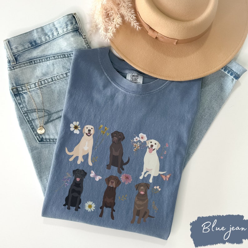 May include: A blue jean t-shirt with a graphic of five Labrador Retrievers in different colors, surrounded by flowers and butterflies. The text "COMFY COLORS" is visible on the shirt.