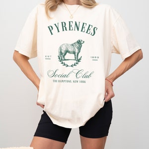 May include: A cream-colored t-shirt with a green graphic of a dog and the text "Pyrenees Social Club The Hamptons, New York" printed on the front.